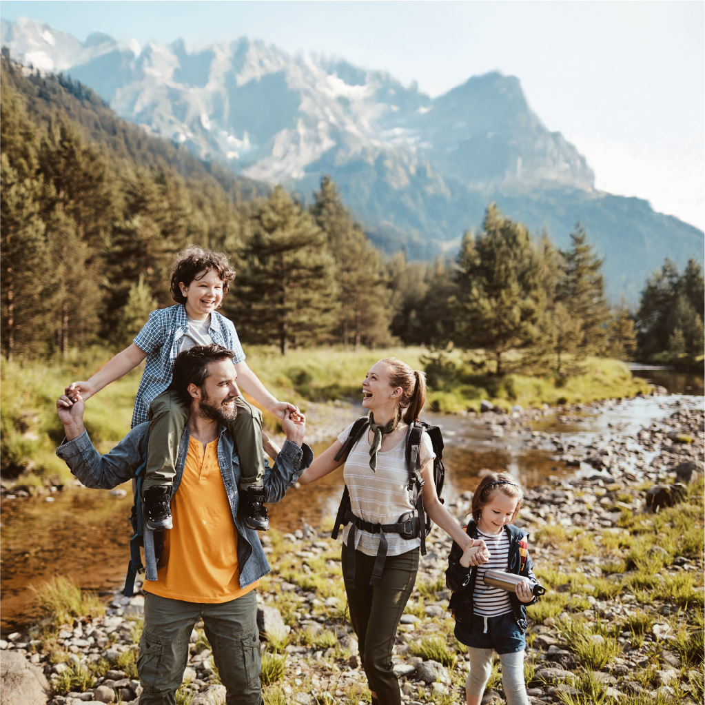 Familie mit zwei jungen Kindern wandert entlang eines Bachlaufes. Im Hintergrund sieht man die Alpen.
